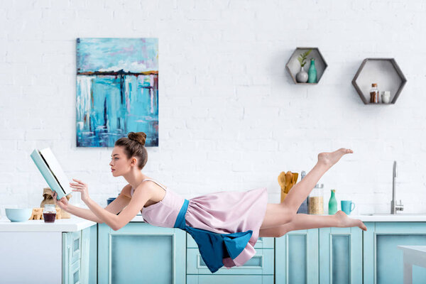 young concentrated barefoot woman levitating in air while reading recipe in cookbook