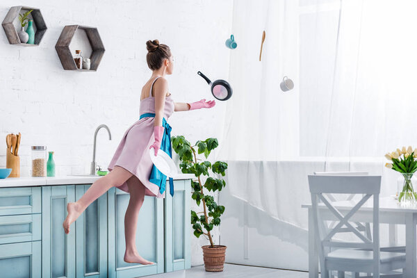 young woman in rubber gloves levitating in air with cooking utensils in kitchen 