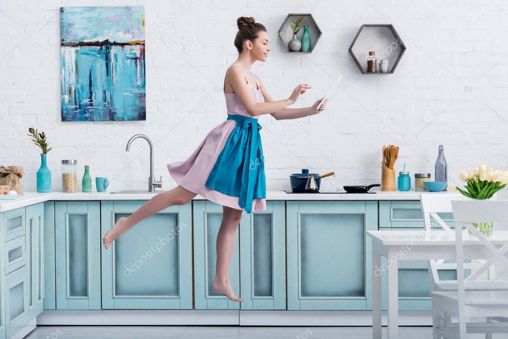 young happy barefoot woman levitating in air while using digital tablet in kitchen