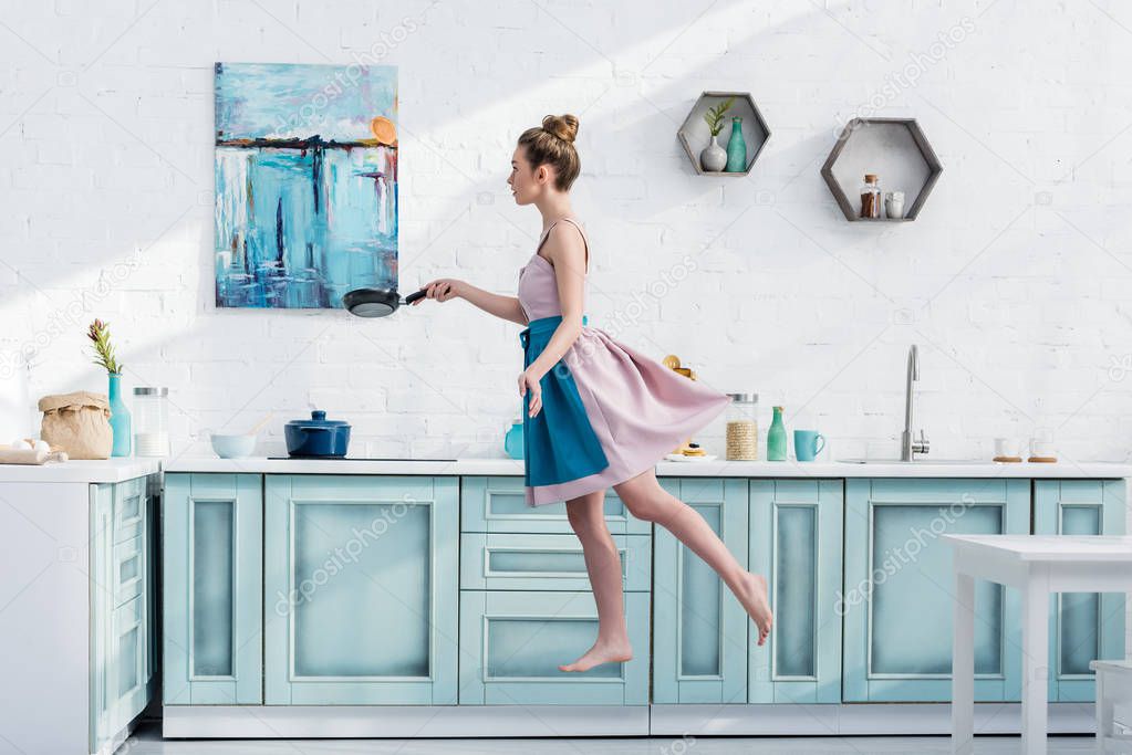 attractive barefoot young woman in apron levitating in kitchen while holding pan 