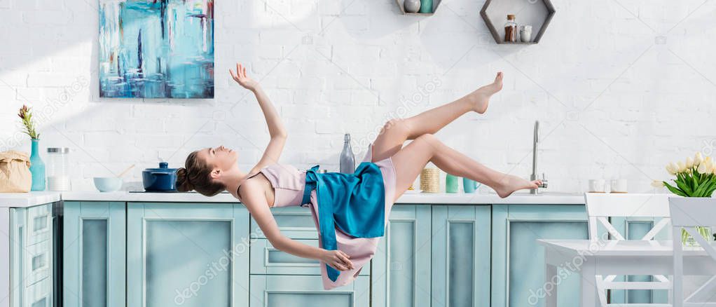 panoramic shot of young woman in apron and dress levitating in air in kitchen