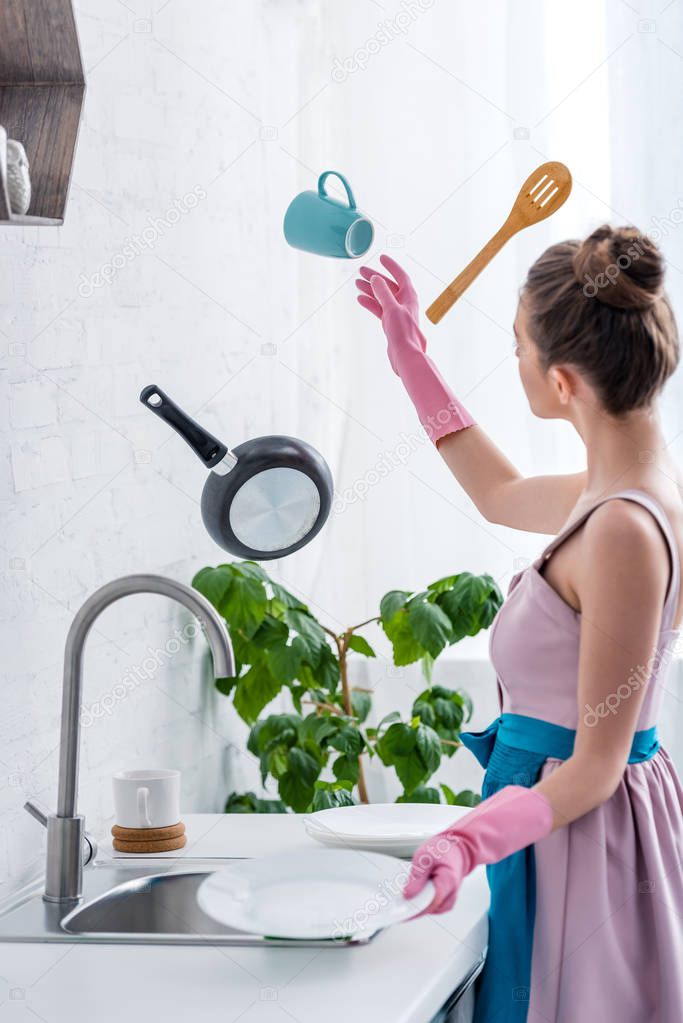young woman in rubber gloves looking at flying in air cooking utensils in kitchen