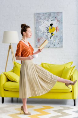 elegant young woman holding book and coffee cup while marching in living room clipart