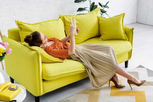 本を読みながら黄色のソファーに横になっているエレガントな若い女性 — ストック写真