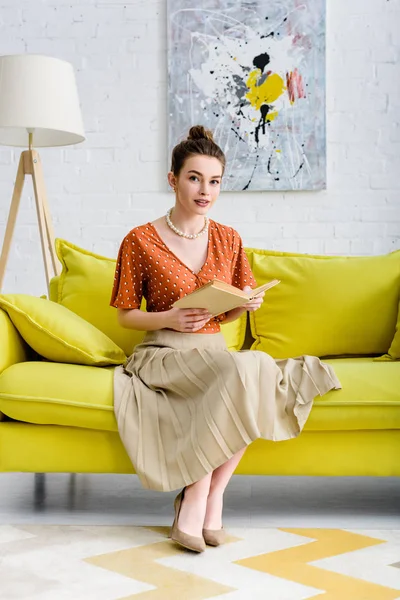 魅力的なエレガントな若い女性の本を押しながらソファに座ってカメラ目線 — ストック写真
