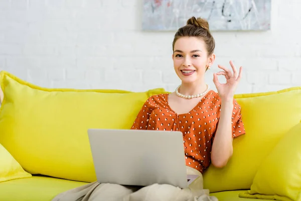 幸せなエレガントな若い女黄色のソファの上に座って 居間でノート パソコンを使用して いい兆しを見せ — ストック写真