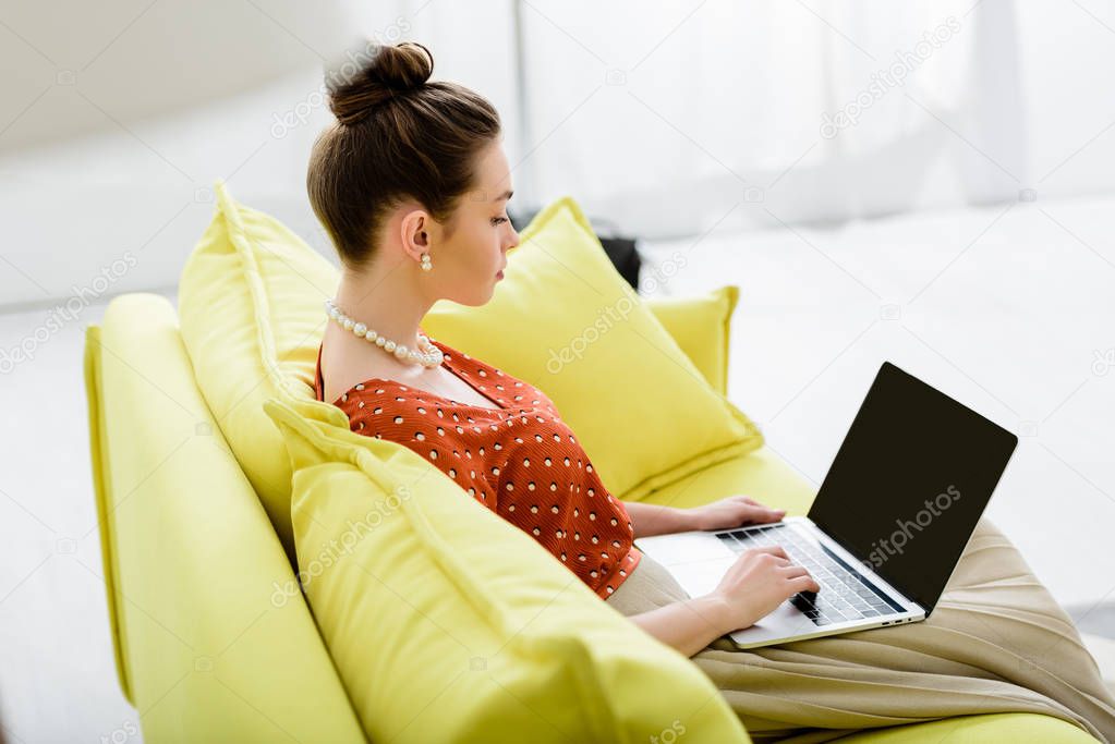 confident fashionable young woman in pearl necklace sitting on yellow sofa and using laptop with blank screen 