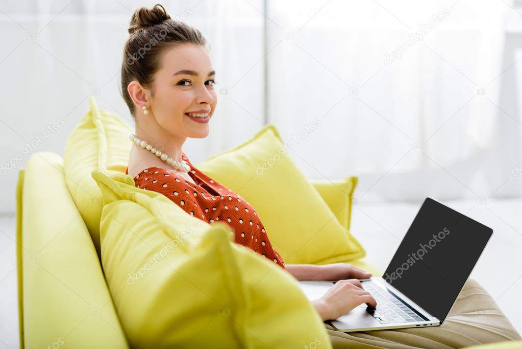 smiling elegant young woman in pearl necklace sitting on yellow sofa and using laptop with blank screen 