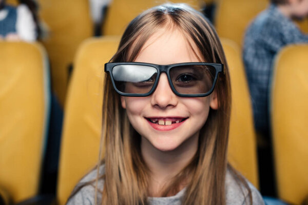 selective focus of smiling child in 3d glasses looking at camera while sitting in cinema