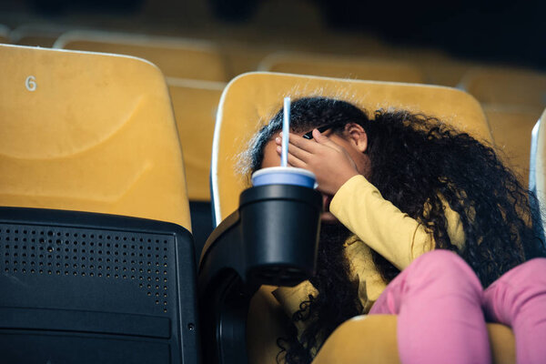 scared african american child holding hand on face while sitting in cinema seat with paper cup in cup holder