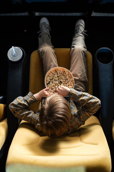 overhead view of boy eating popcorn while sitting in cinema chair near paper cup