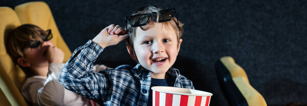 panoramic shot of cute boy looking at camera and holding stripped paper cup with pop corn