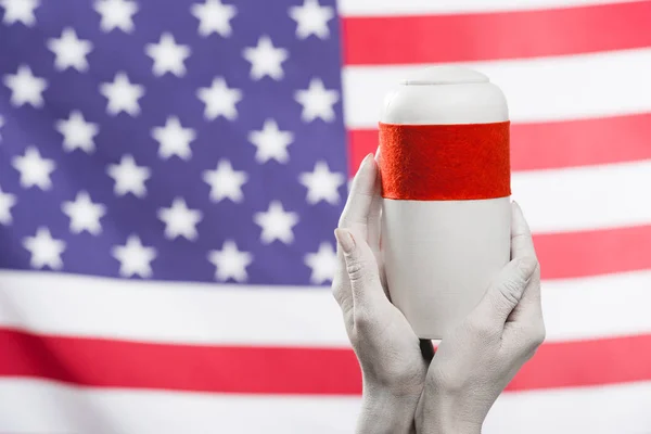 cropped view of female hands painted in white holding funeral urn near american flag