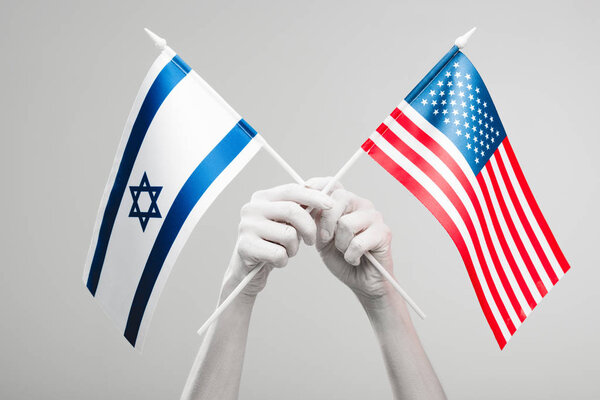 cropped view of female hands painted in white holding crossed american and israel flags isolated on grey 