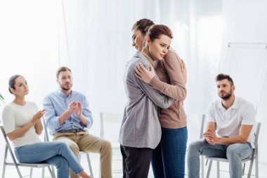women hugging while group of people sitting and applauding during therapy session clipart