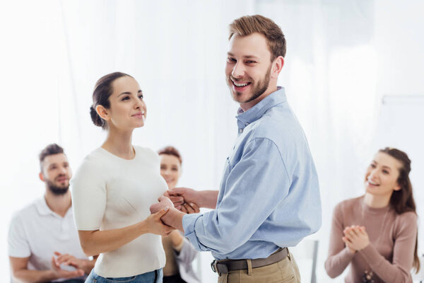 улыбающийся мужчина смотрит в камеру и держит женщину за руку во время сеанса групповой терапии
