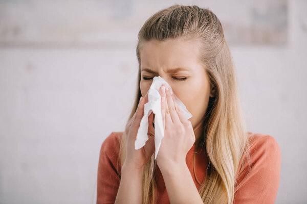 blonde woman sneezing while holding tissue near nose 