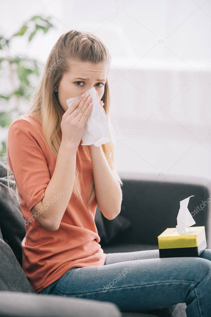 blonde woman sneezing while sitting on sofa and looking at camera 