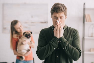 selective focus of man sneezing in tissue with closed eyes while standing near woman with dog  clipart
