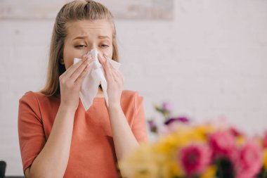 selective focus of blonde woman looking at flowers and sneezing in tissue while having pollen allergy clipart