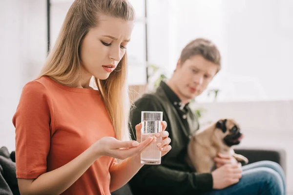 錠剤とパグの男の近くに水のガラスを保持している犬にアレルギーがある動揺の金髪女性の選択と集中 — ストック写真