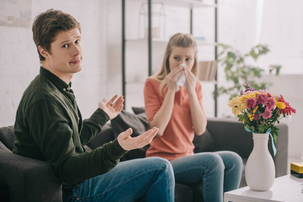 handsome man showing shrug gesture while sitting near vase with flowers and sneezing girl with pollen allergy