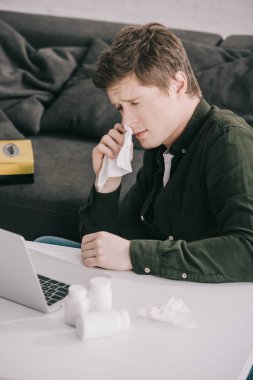 upset man holding tissue while sitting with closed eyes near bottles with pills and laptop  clipart