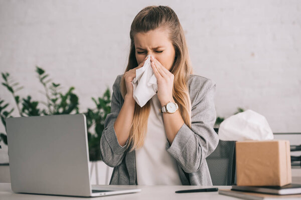 blonde businesswoman sneezing in tissue near laptop in office 