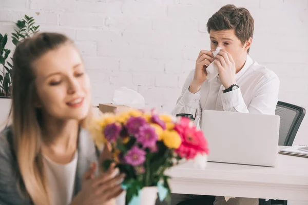 selective focus of sneezing man with pollen allergy near blonde coworker looking at flowers in vase