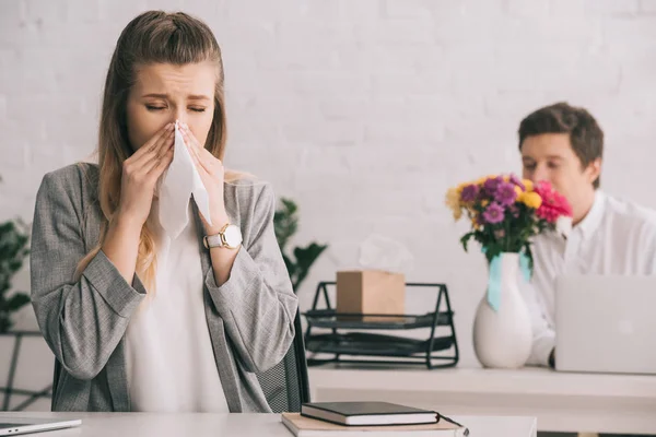 blonde businesswoman with pollen allergy sneezing in tissue near coworker smelling flowers in office