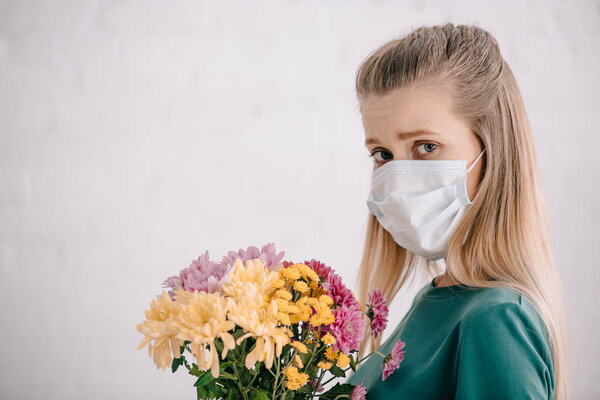 blonde woman with pollen allergy wearing medical mask and holding flowers while looking at camera 
