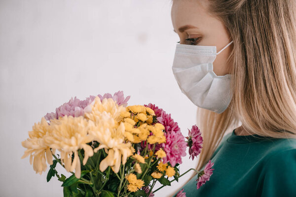 blonde woman with pollen allergy wearing medical mask and looking at bouquet of flowers 
