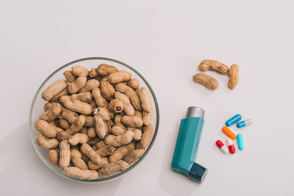 top view of pills near blue inhaler and glass bowl with tasty peanuts on grey 