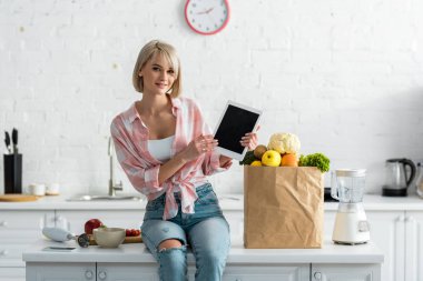 beautiful blonde woman holding digital tablet with blank screen near paper bag with ingredients   clipart