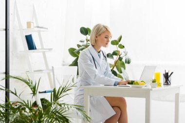 attractive blonde nutritionist using laptop near plate with organic food clipart