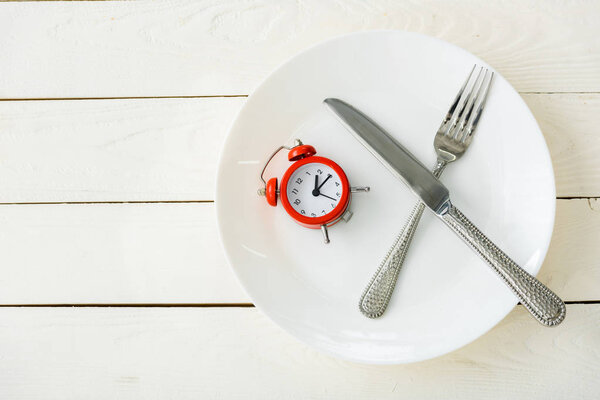top view of white plate with cutlery and red alarm clock on wooden surface 
