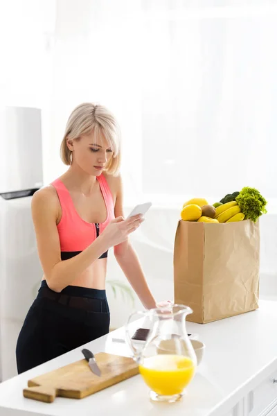 Blonde Vrouw Sportkleding Kijken Naar Smartphone Buurt Papieren Zak Met — Stockfoto