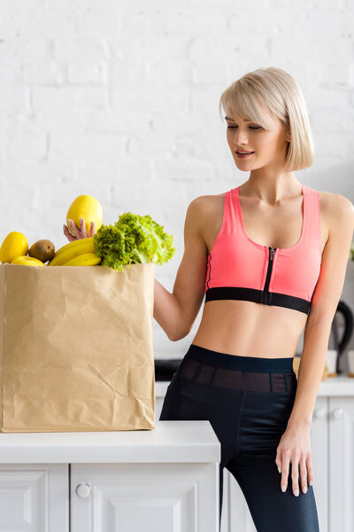 cheerful blonde woman in sportswear looking at paper bag with groceries