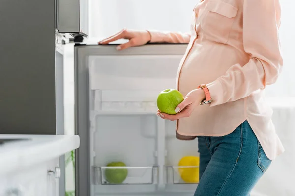 台所で冷蔵庫の近くに緑のリンゴを保持する妊婦のクロップビュー — ストック写真