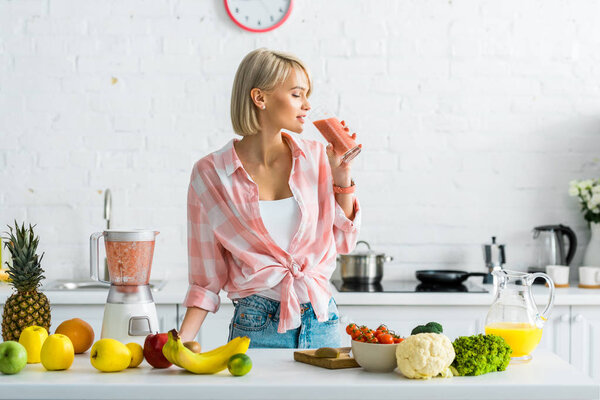 attractive blonde woman drinking tasty smoothie near ingredients in kitchen 