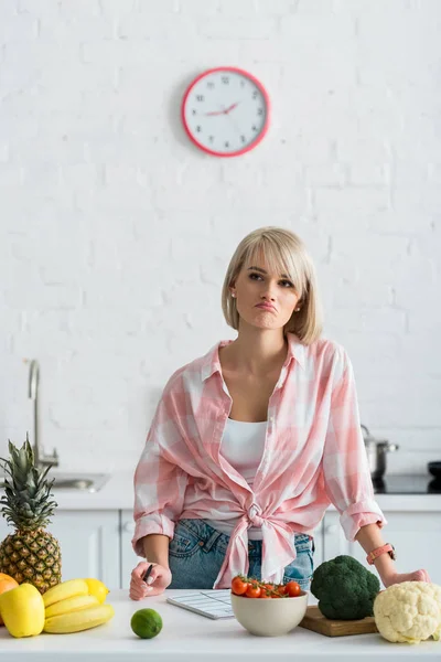 sad blonde woman holding pen near notebook and ingredients in kitchen