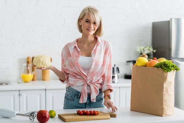 cheerful blonde girl holding cauliflower near paper bag with groceries
