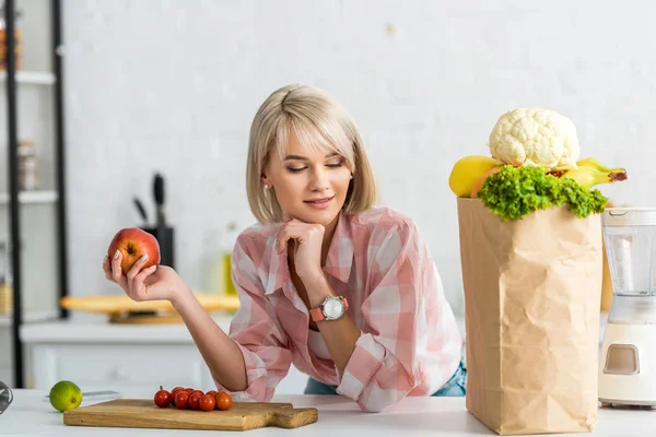 Boldog Szőke Kezében Alma Miközben Nézi Papírzacskót Élelmiszert Konyhában — Stock Fotó