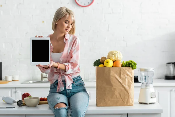 食材と紙袋の近くに空白の画面でデジタルタブレットを保持している魅力的なブロンドの女性 — ストック写真