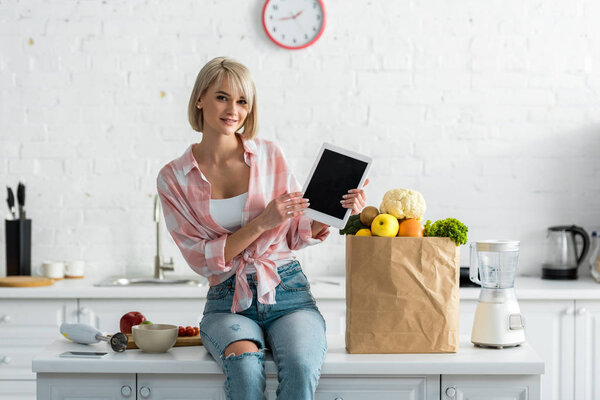 beautiful blonde woman holding digital tablet with blank screen near paper bag with ingredients  