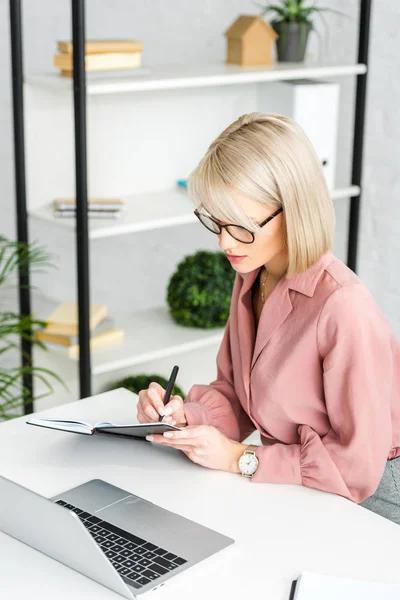Pensive Young Blonde Woman Glasses Writing Notebook While Sitting Laptop — Stock Photo, Image