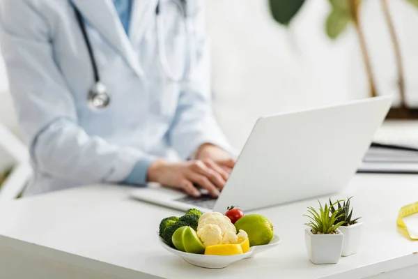 有機食品とプレートの近くにノートパソコンを使用して栄養士のクロップビュー — ストック写真