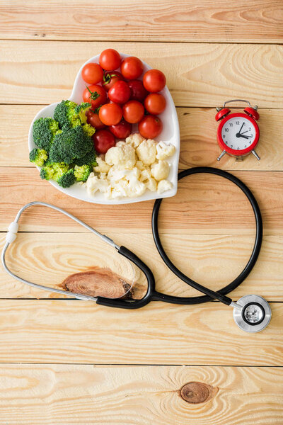 top view of vegetables on heart-shape plate near stethoscope and retro alarm clock on wooden surface 