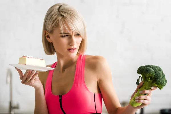 Blonde Woman Looking Green Broccoli While Holding Saucer Sweet Cake — Fotografie, imagine de stoc