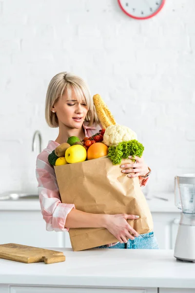 Привлекательная блондинка держит тяжелый бумажный пакет с продуктами — стоковое изображение Привлекательная Блондинка Держит Тяжелый Бумажный Пакет Продуктами Стоковая Картинка